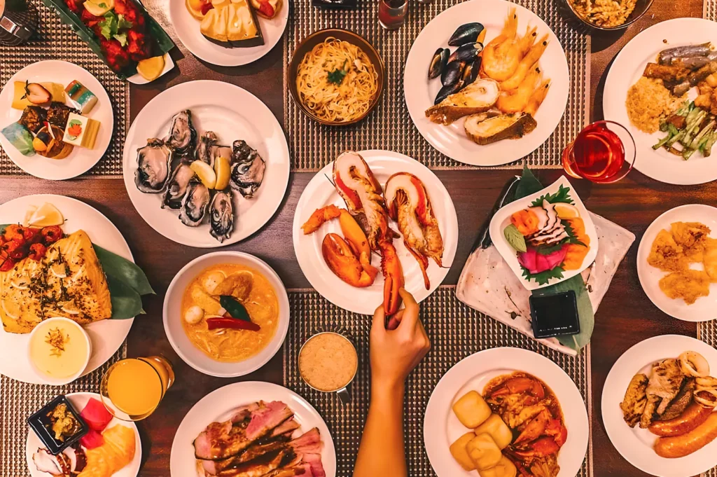 A variety of gourmet dishes are spread across a table, including seafood, pasta, and sushi. A hand reaches for lobster, creating a lively dining scene.