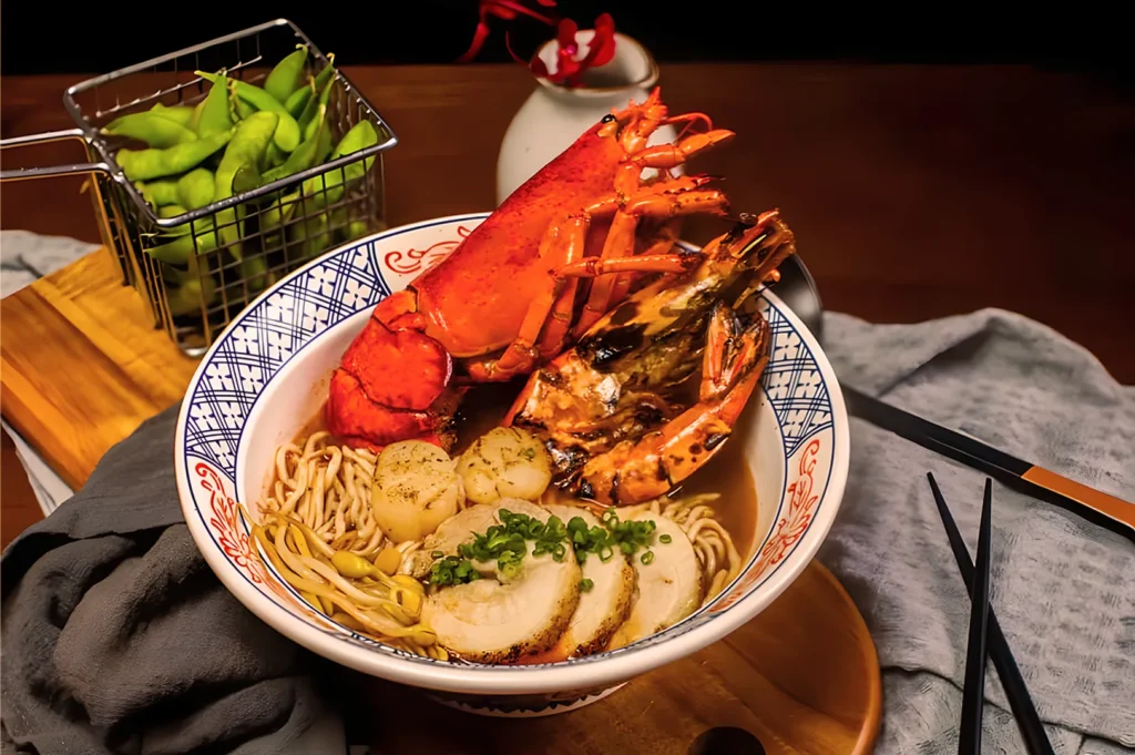 A vibrant bowl of ramen topped with lobster, scallops, and sliced pork in rich broth. Edamame sits in a basket beside black chopsticks, creating a gourmet scene.