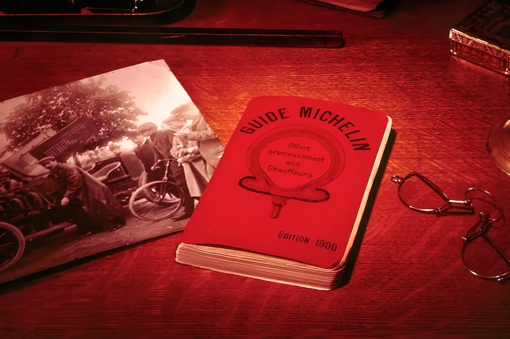A vintage red Michelin Guide from 1900 lies on a wooden table. Beside it, an old sepia photo of people with a vintage car and a pair of reading glasses convey nostalgia.