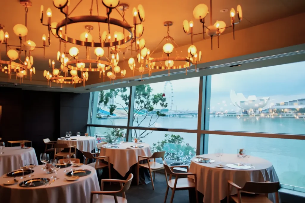 Elegant restaurant with round tables, white tablecloths, and warm chandeliers. Large windows reveal a scenic waterfront and a Ferris wheel.
