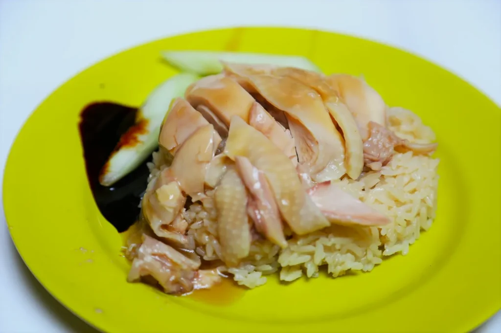 Sliced poached chicken on fragrant rice served on a yellow plate, garnished with cucumber slices and dark soy sauce, conveying a simple, delicious meal.