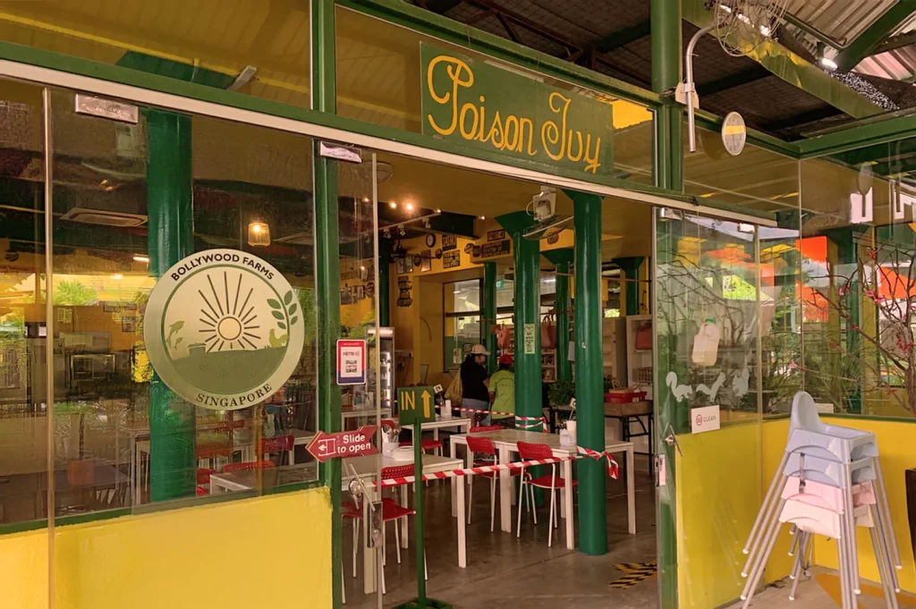 Exterior of "Poison Ivy" café at Bollywood Farms, Singapore. Glass doors open to a cozy interior with red chairs and green pillars, conveying a welcoming atmosphere.