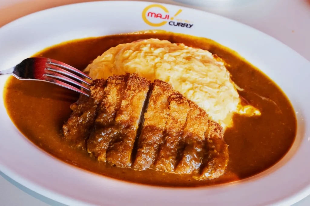 Plate with Japanese curry featuring golden fried cutlet and creamy omelette over rice, topped with rich brown sauce, conveying a warm, hearty meal.