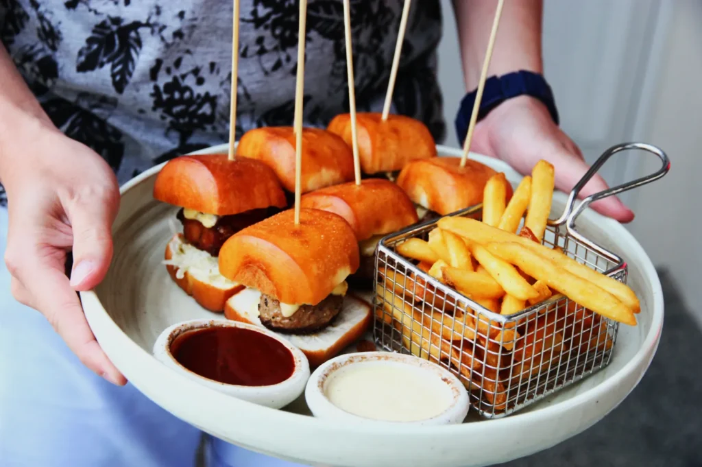 A person holds a white plate with mini burgers on skewers, a metal basket of fries, and two dipping sauces, creating a casual dining vibe.