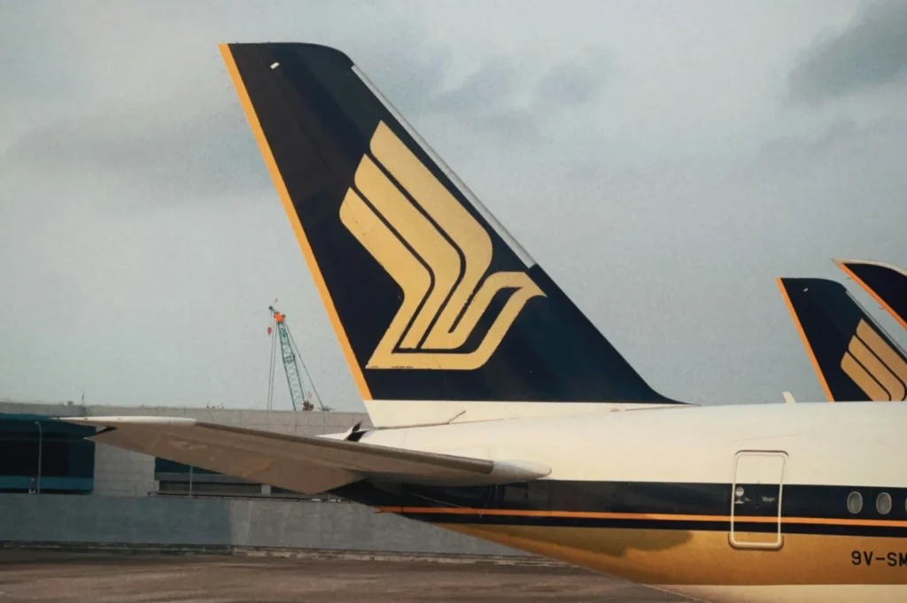 The image shows the tail fin of an airplane with a stylized yellow bird logo against a navy background, parked at an airport under a cloudy sky.