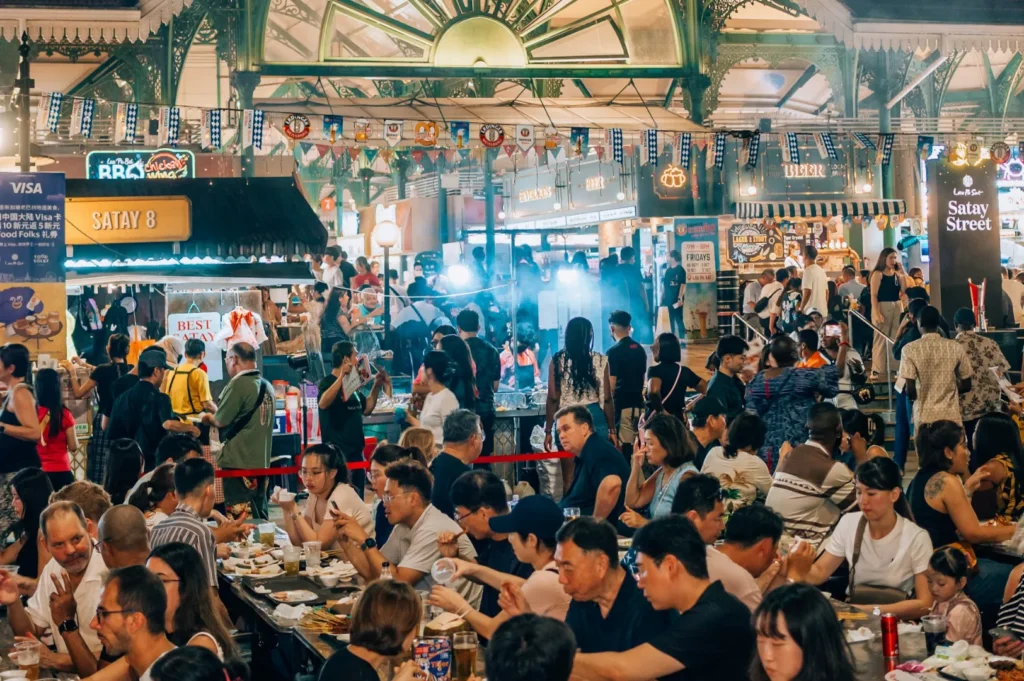 A bustling night market filled with people enjoying food at various stalls. Colorful lights and banners hang above, creating a lively and festive atmosphere.