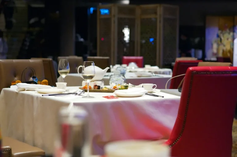 Elegant restaurant setting with white tablecloths, red cushioned chairs, and neatly arranged tableware. Soft lighting creates a warm, inviting ambiance.