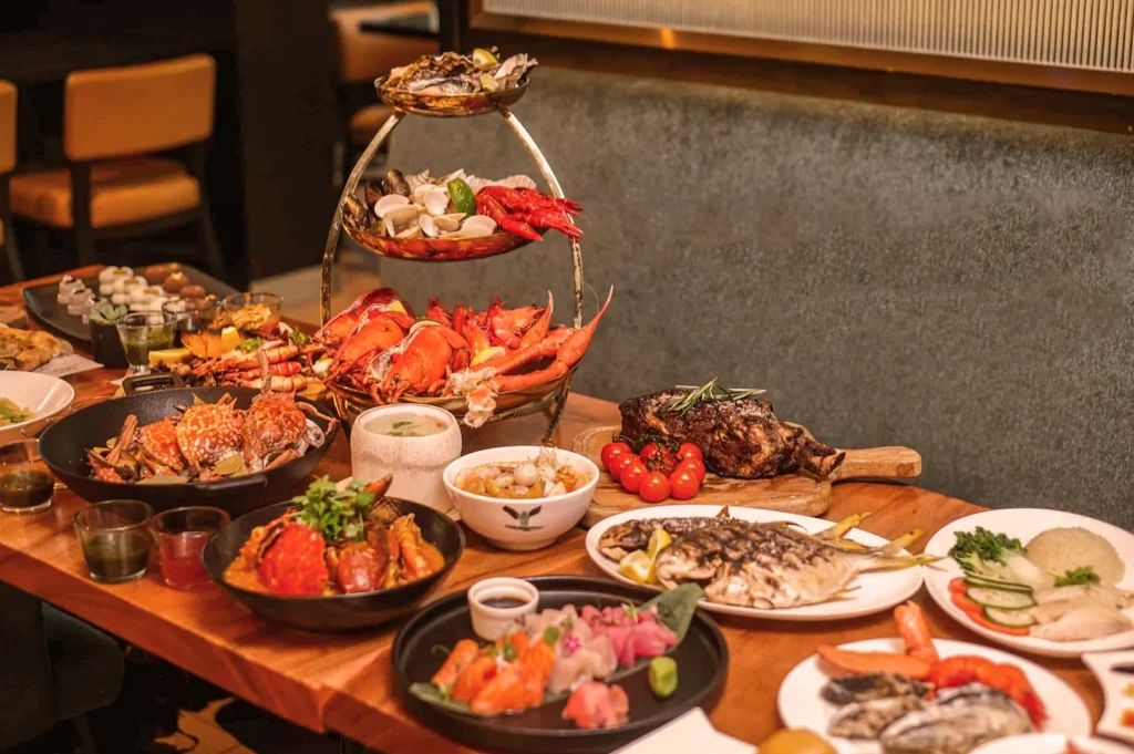 A lavish seafood spread on a wooden table includes lobster, crab, oysters, sushi, grilled fish, and steak, conveying an indulgent and opulent dining experience.