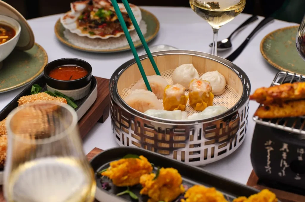 A bamboo steamer filled with assorted dim sum sits on a dining table surrounded by plates of appetizers, a dipping sauce, and glasses of white wine.