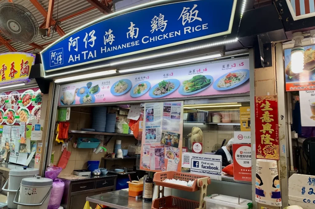 A vibrant food stall with a blue sign reading "Ah Tai Hainanese Chicken Rice" in a bustling market. Colorful menu photos depict various dishes. The scene is lively and inviting.
