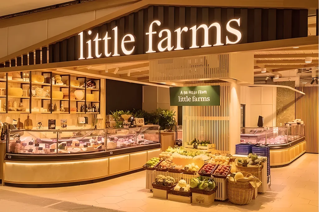 Warmly lit grocery store entrance labeled "little farms," featuring fresh produce in baskets and a deli counter, creating an inviting, fresh atmosphere.