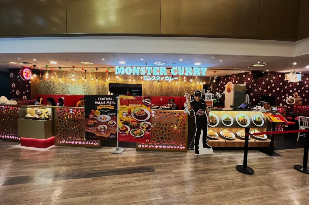Restaurant storefront with "Monster Curry" sign. Display features vibrant meal images and promotional posters. Warm lighting and casual atmosphere.