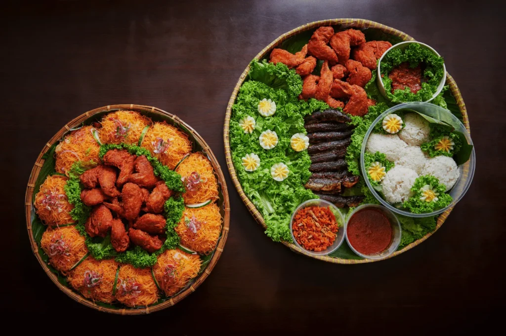 Two woven trays on a dark table, filled with various dishes. One tray holds fried chicken and shredded toppings; the other features rice, grilled meat, and garnishes on lettuce.
