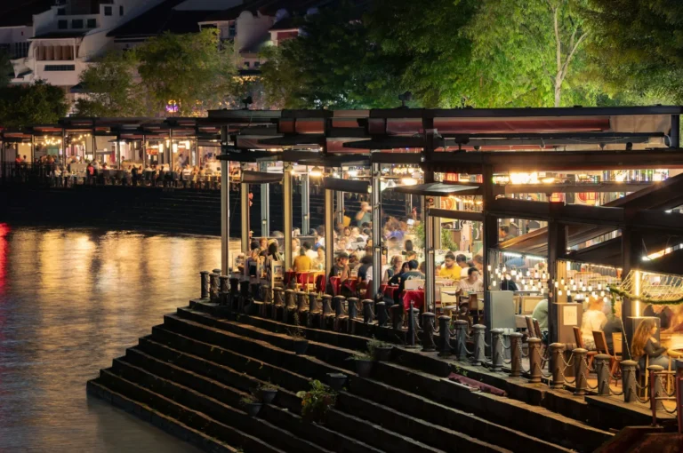 Busy riverside restaurant at night with warm lights and reflections on water, bustling with diners. Trees and adjacent eateries line the background.