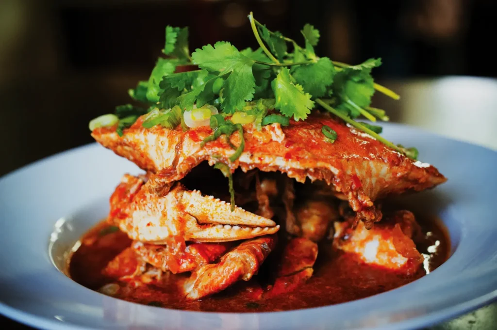 A vibrant dish of spicy crab in a thick red sauce is garnished with fresh cilantro. The crab is served in a white bowl, evoking richness and warmth.