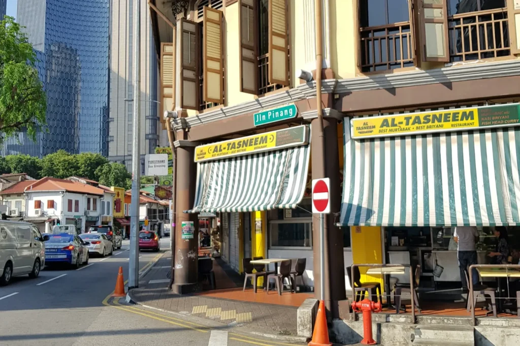 Street scene with a corner building hosting "Al-Tasneem" restaurant. Striped awnings, open shutters, cars, and skyscrapers create a vibrant urban atmosphere.