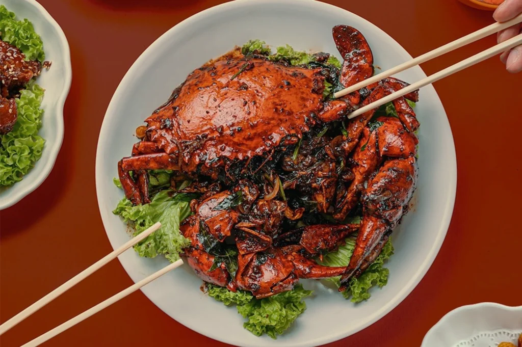A vibrant red, glossy, cooked crab on a white plate, garnished with fresh green lettuce. Chopsticks are poised to pick it up, conveying a sense of anticipation.