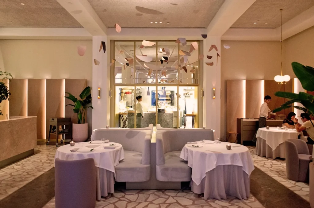 Elegant restaurant interior with soft lighting, neutral tones, and plush seating. Tables are covered with white cloths, creating a sophisticated ambiance.