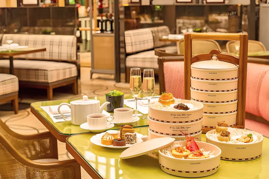 Elegant afternoon tea setting with tiered trays of pastries and savory bites on a green table. Includes teapot, cups, and two champagne flutes.