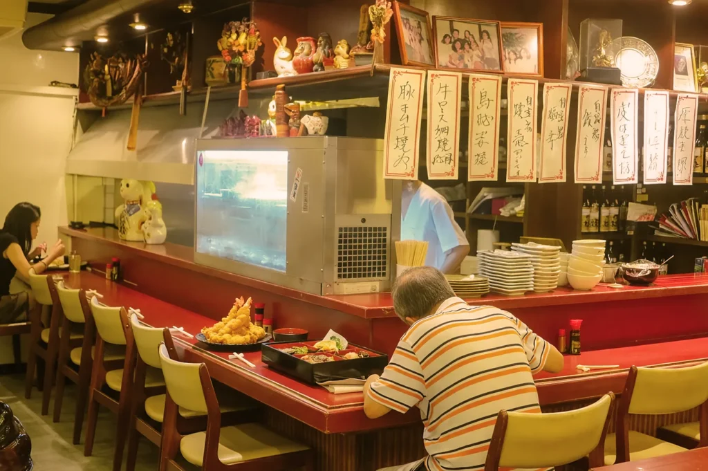Cozy Japanese restaurant interior with wooden decor. Two patrons sit at a red counter enjoying their meals; dishes and decorative ornaments are displayed. Calm atmosphere.