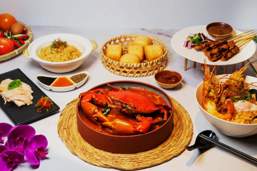 A vibrant spread of Asian cuisine featuring a large dish of chili crab at the center, surrounded by rice, satay skewers, seafood noodles, and steamed buns.