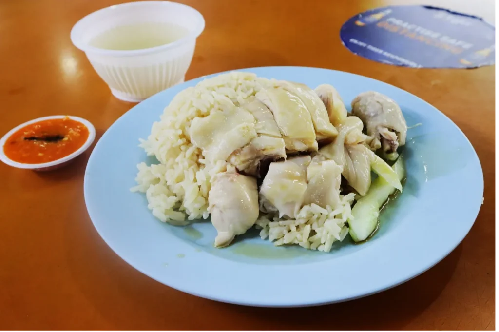 A plate of Hainanese chicken rice with tender chicken slices over fragrant rice, garnished with cucumber. Accompanied by a bowl of clear soup and chili sauce on a wooden table.