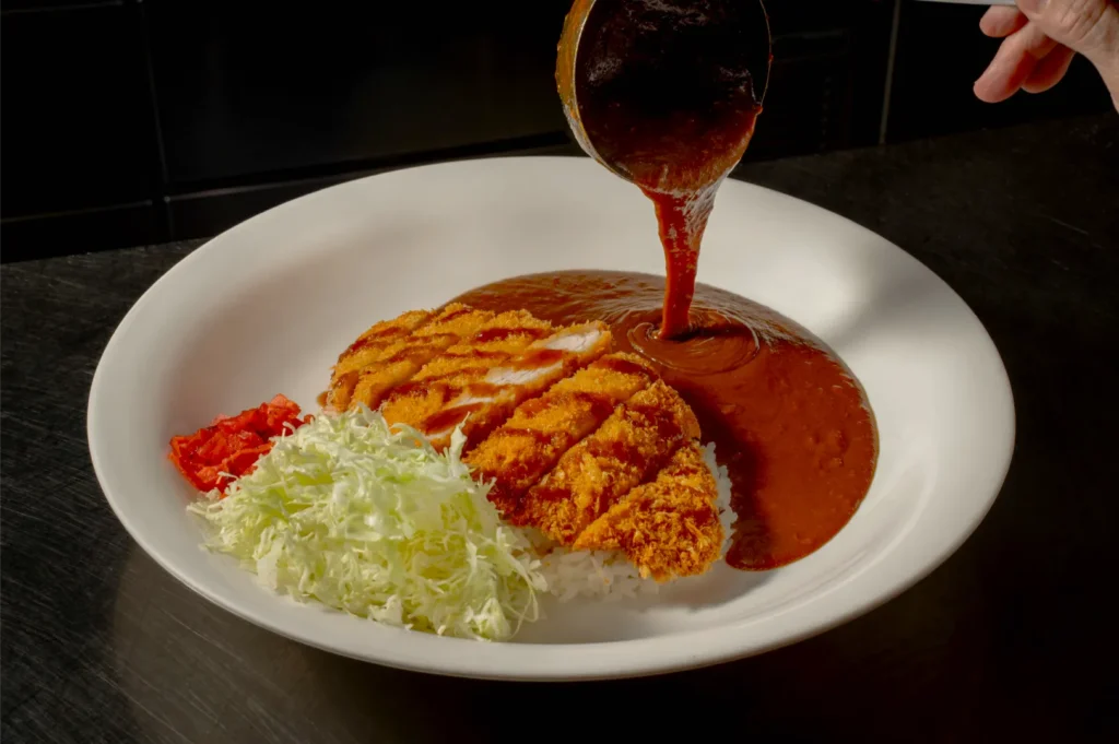 A white plate with crispy breaded chicken cutlet on rice, drizzled with dark curry sauce from a ladle. Shredded cabbage and red pickles on the side.