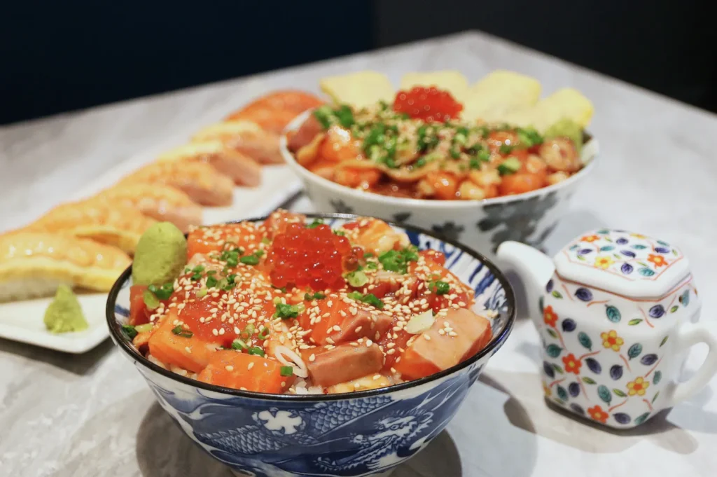 Two bowls of vibrant seafood donburi topped with salmon roe, sesame seeds, and green onions. A floral teapot and sushi rolls are in the background.