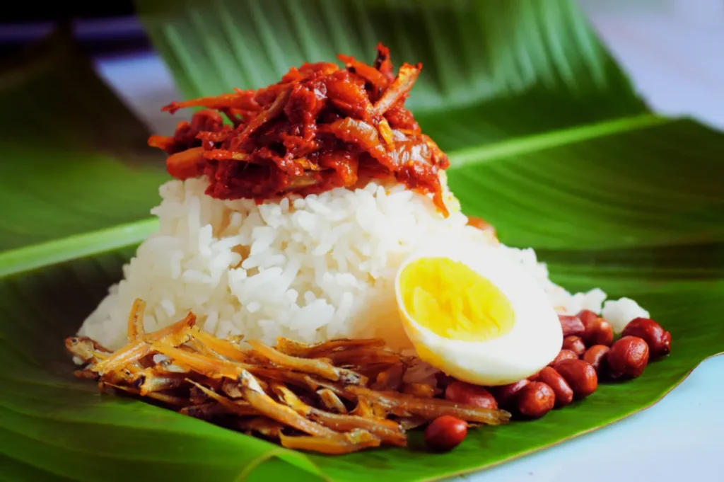 A traditional dish of nasi lemak on banana leaves, featuring white rice, sambal, a halved boiled egg, fried anchovies, and roasted peanuts.
