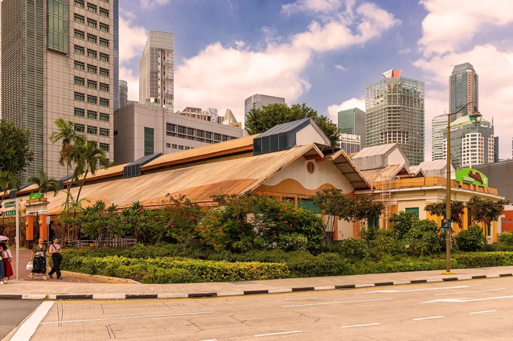 A rustic, traditional-style building with a sloped roof is surrounded by greenery, set against a backdrop of modern skyscrapers under a blue, cloudy sky.