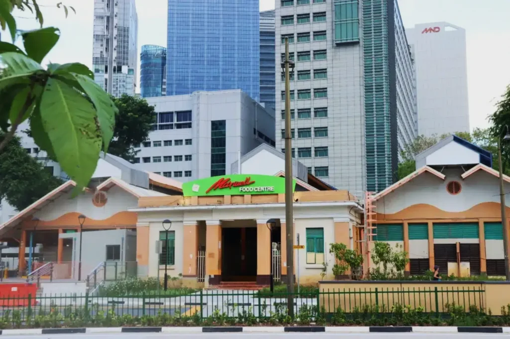 Historic building of Maxwell Food Centre with beige facade and green sign, set against modern high-rise buildings. Lush greenery in foreground.