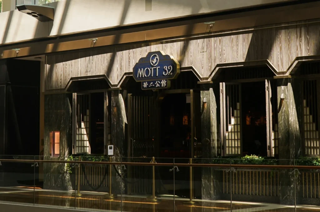 Exterior of an upscale restaurant with wooden paneling and a sign reading "Mott 32." Subtle lighting creates a sophisticated, inviting atmosphere.
