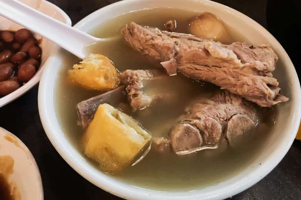 Bowl of soup with pork ribs, yam, and taro chunks. A spoon rests on the bowl's edge. Side dishes of peanuts and soy sauce are partially visible.