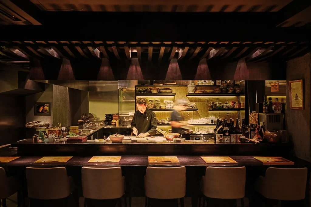 A warmly lit open-kitchen restaurant with a chef preparing food, surrounded by shelves of pottery and bottles. Bar stools face the counter, creating an intimate, welcoming atmosphere.