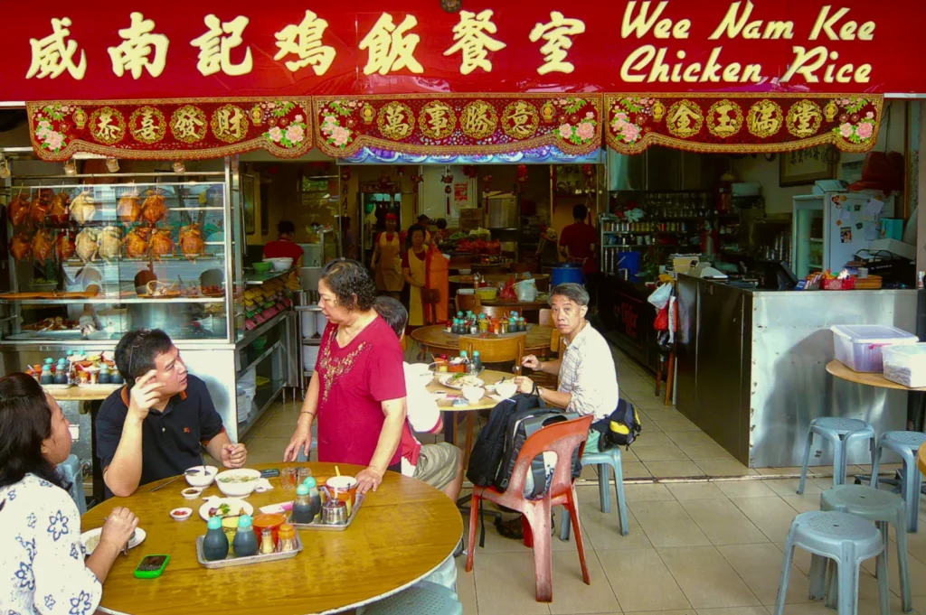A bustling chicken rice shop with red signage, featuring customers seated at round tables, engaging in conversation and enjoying food. The atmosphere is lively and casual.