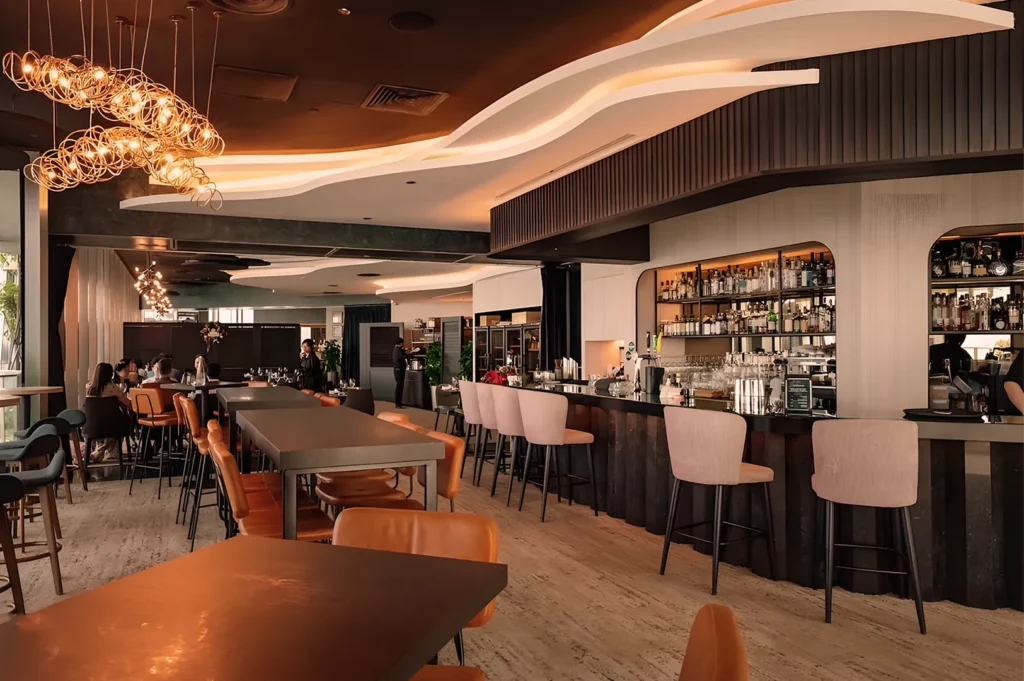 Modern restaurant interior with a warm, inviting ambiance. Features sleek bar counter, elegant lighting, and stylish seating. Soft, atmospheric lighting.