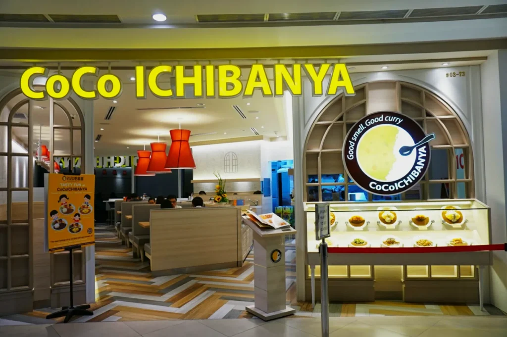 Front view of CoCo ICHIBANYA restaurant with a bright sign. Inside are red lamps, cozy booths, and a welcoming ambiance. A display shows curry dishes.