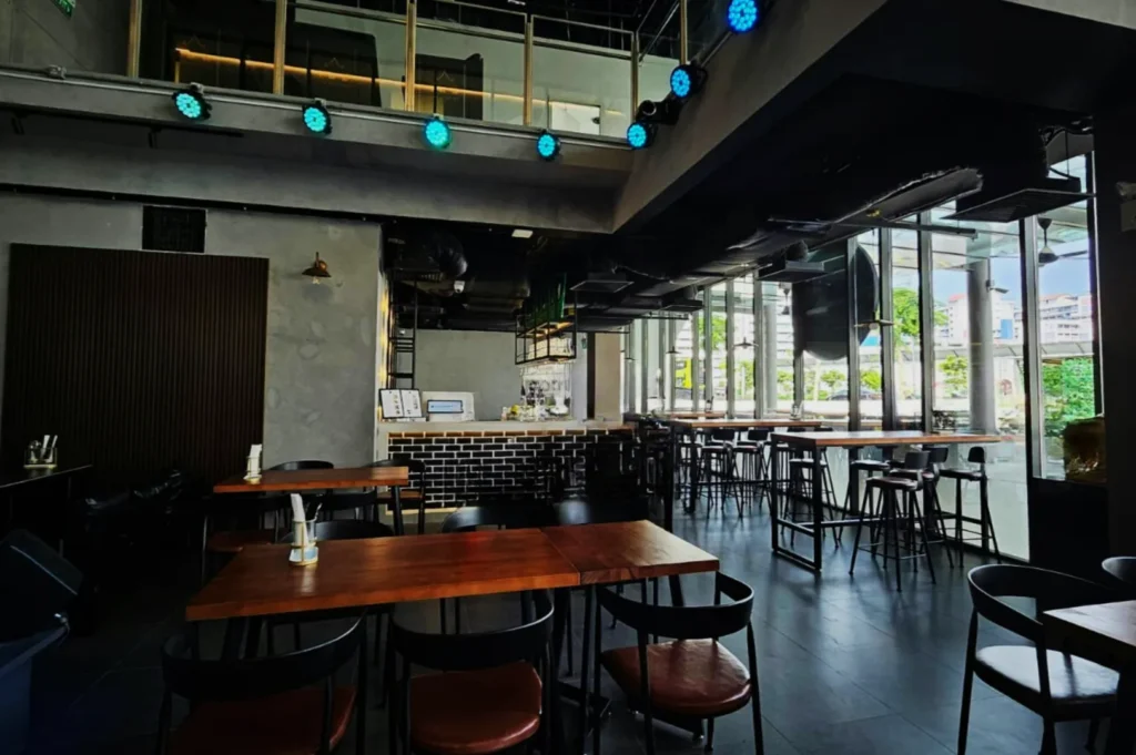 Spacious modern cafe interior with wooden tables, black chairs, floor-to-ceiling windows, and a brick counter. Dim lighting creates a cozy atmosphere.
