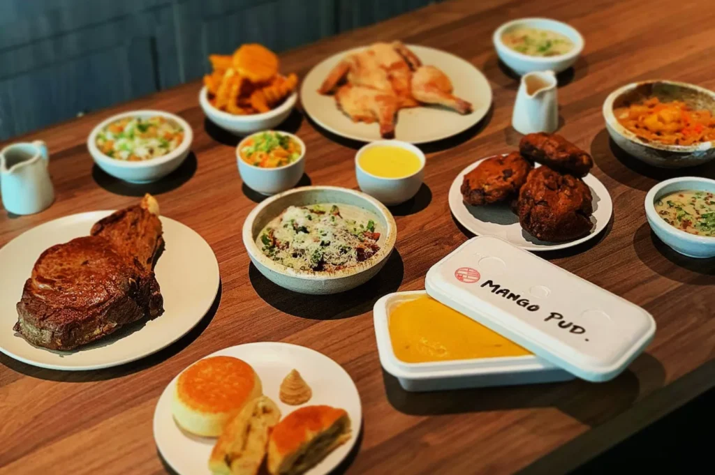 A wooden table is filled with a variety of dishes: a large steak, rotisserie chicken, biscuits, bowls of salad, side dishes, and mango pudding. The food is arranged neatly, creating an inviting and hearty atmosphere.
