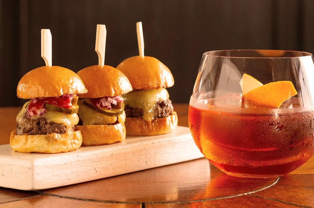 Three mini cheeseburgers on a wooden board, topped with onions, pickles, and ketchup, paired with a glass of amber cocktail garnished with an orange peel.