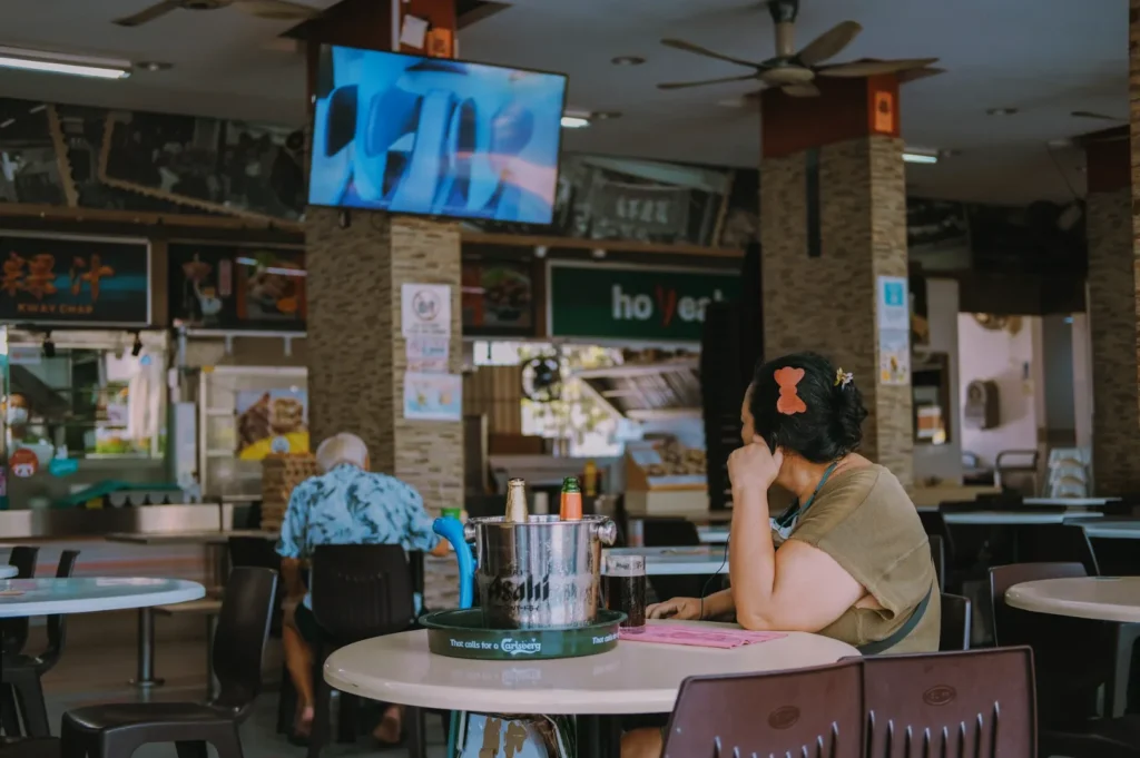 Man seated alone in an airy food court, looking up at a TV. The area is calm, with empty tables and a bustling stall in the background.