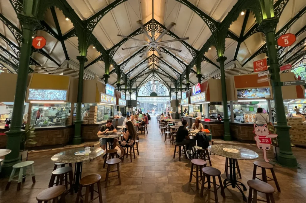 Busy indoor food market with ornate iron columns and high ceilings. People enjoy meals at round tables. Bright food stalls line the sides, creating a lively atmosphere.