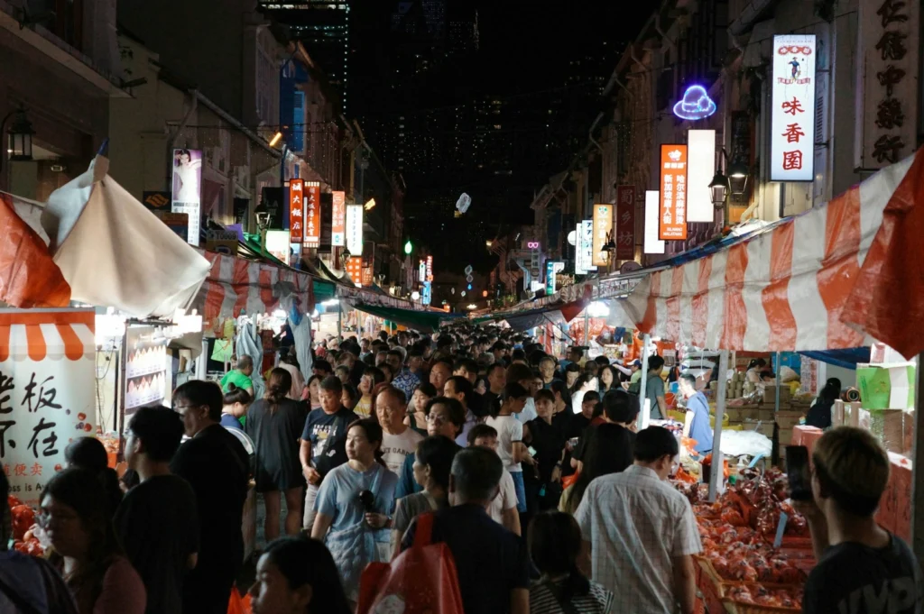 Busy night market scene with crowds of people navigating between rows of brightly lit stalls under striped canopies, creating a lively, bustling atmosphere.