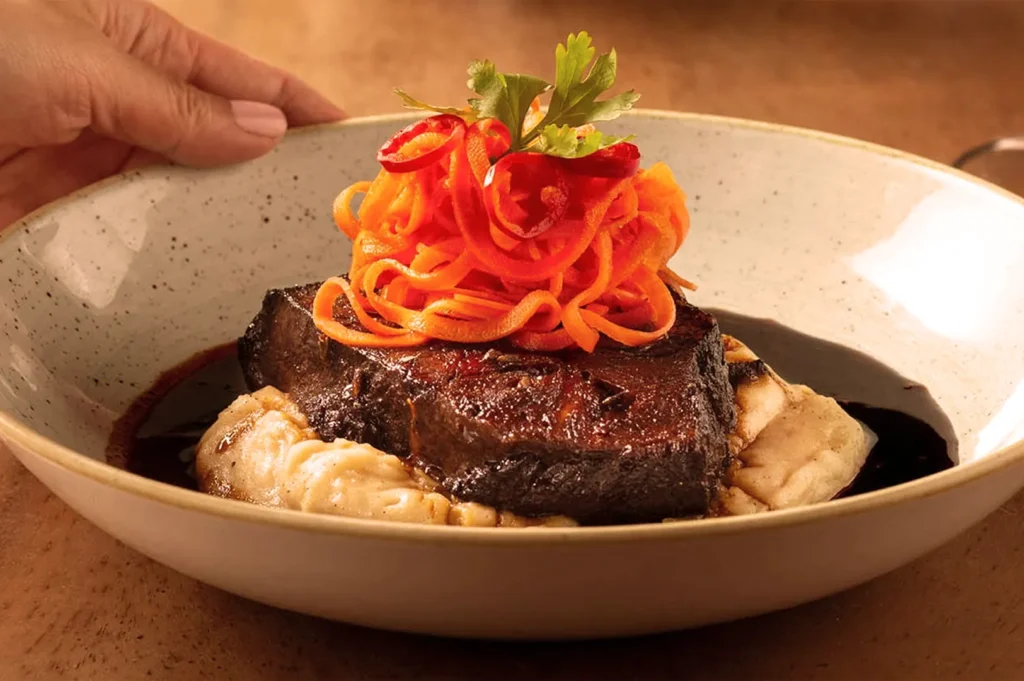 A gourmet dish featuring a seared meat slice atop creamy puree, garnished with spiralized carrots and a sprig of parsley, in an earthy-toned bowl.