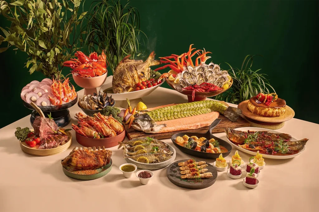 A lavish seafood spread on a table with lobster, crab, oysters, whole fish, and prawns. The background features lush green plants, creating an elegant and rich atmosphere.