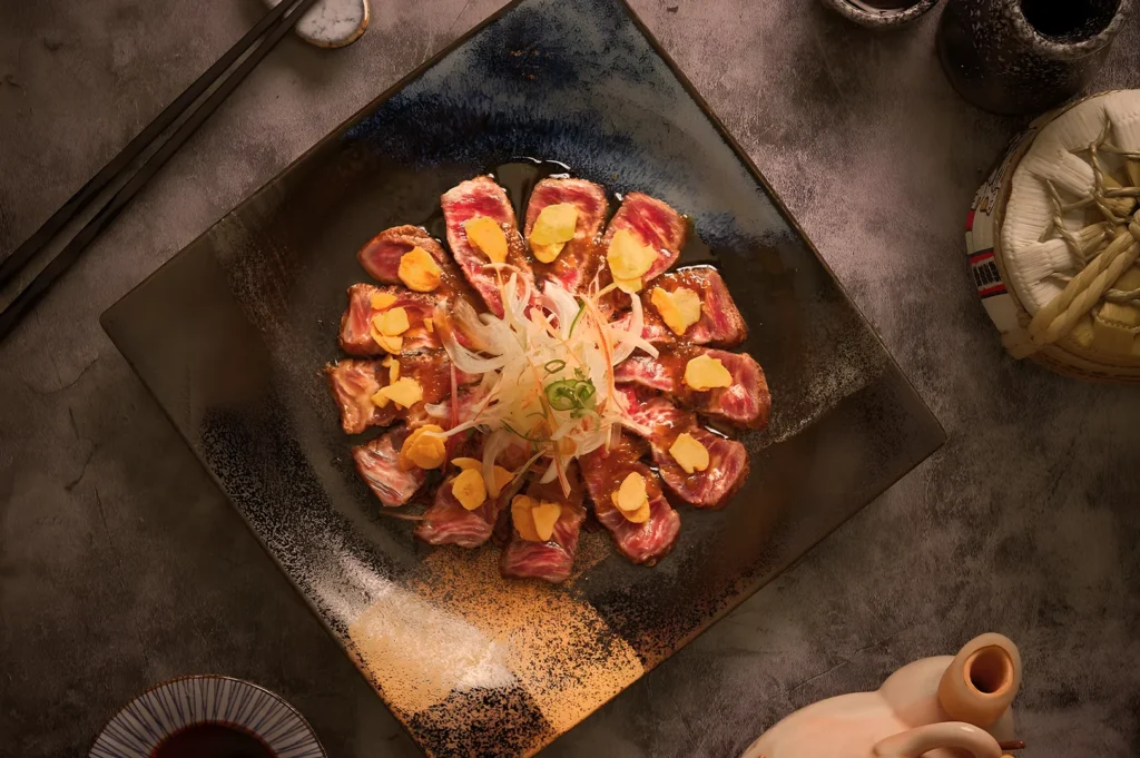 A beautifully arranged dish features thinly sliced, marbled beef topped with sauces and garnishes, surrounded by delicate shaved onions on a textured dark plate.