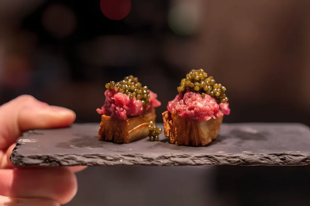 A hand holds a black slate with two small bruschetta topped with raw minced meat and green caviar. The setting is dimly lit, creating a gourmet ambiance.