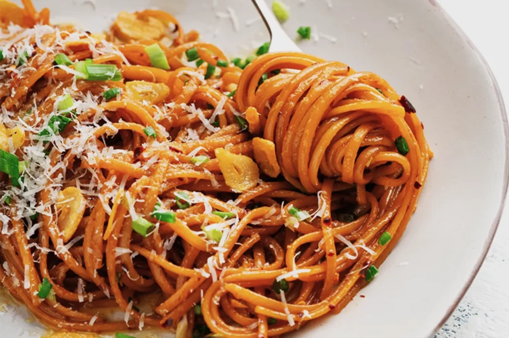 A plate of spaghetti aglio e olio with sliced garlic, chopped green herbs, and grated cheese, with a fork entwined in noodles, conveying warmth.