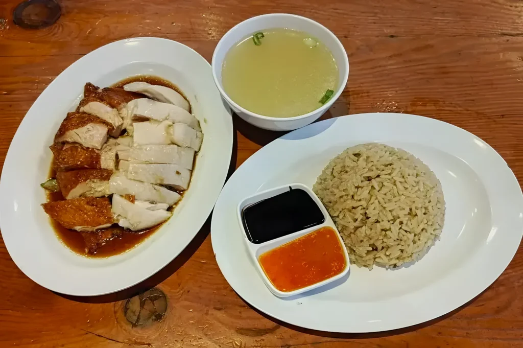 Plate of sliced roasted chicken with soy, side of rice, broth soup with scallions, on a wooden table. Includes black and orange dipping sauces.