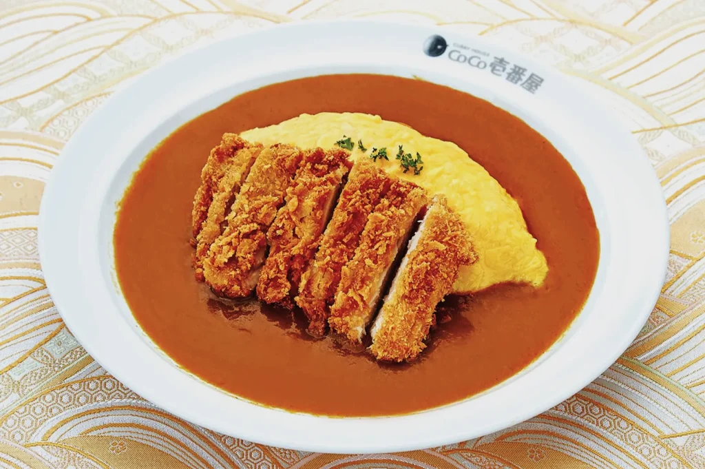 A plate of Japanese curry rice with sliced crispy katsu and a soft omelette on top. The dish is vibrant and inviting, set on a patterned tablecloth.
